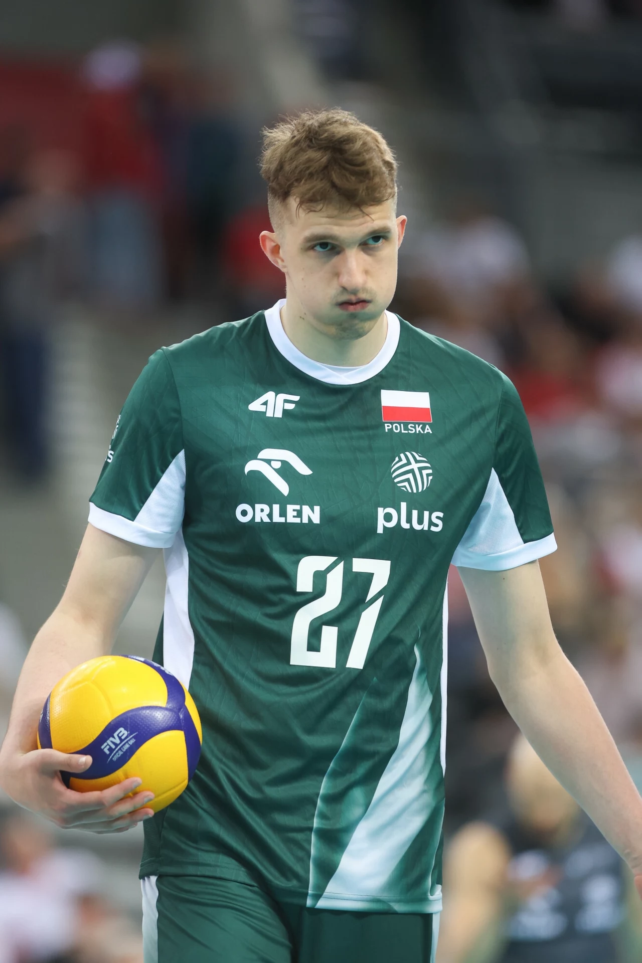 Michał Gierżot Młody siatkarz ubrany w zieloną koszulkę reprezentacji Polski z numerem 27 stoi na boisku z piłką do siatkówki w dłoni, skupiony, gotowy do wykonania zagrywki, na tle rozmytej publiczności.