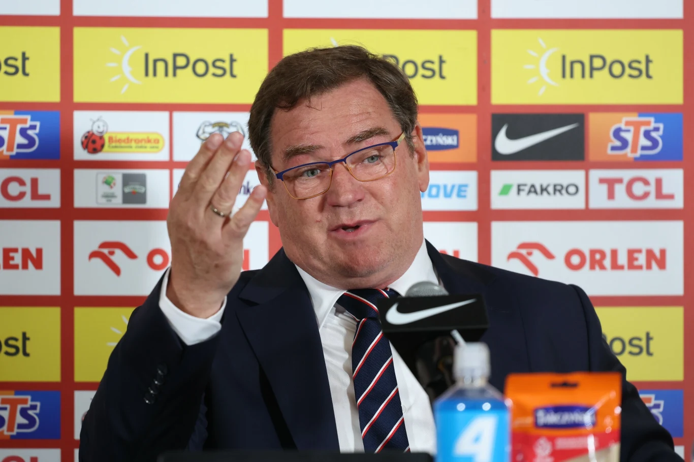 Jan Urban podczas konferencji prasowej Mężczyzna w garniturze i okularach wykonuje gest dłonią podczas konferencji prasowej, w tle widoczne logotypy sponsorów, na pierwszym planie mikrofon oraz butelka z wodą i opakowanie z napojem.