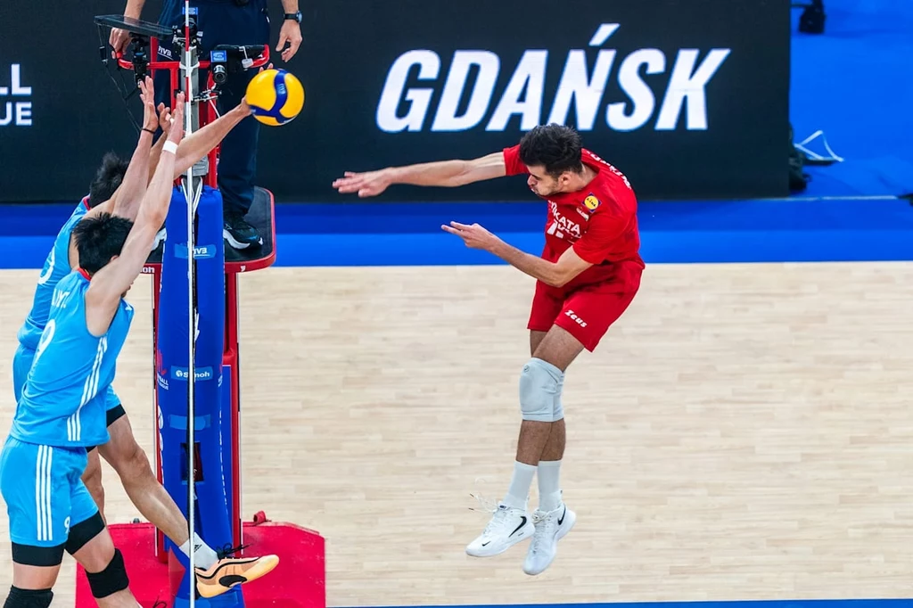Liga Narodów siatkarzy. Kadr z meczu Chiny - Bułgaria Dynamiczna akcja podczas meczu siatkówki, zawodnik w czerwonym stroju z wyskoku atakuje piłkę przez siatkę, a dwóch graczy drużyny przeciwnej próbuje ją zablokować; w tle widoczny napis Gdańsk.
