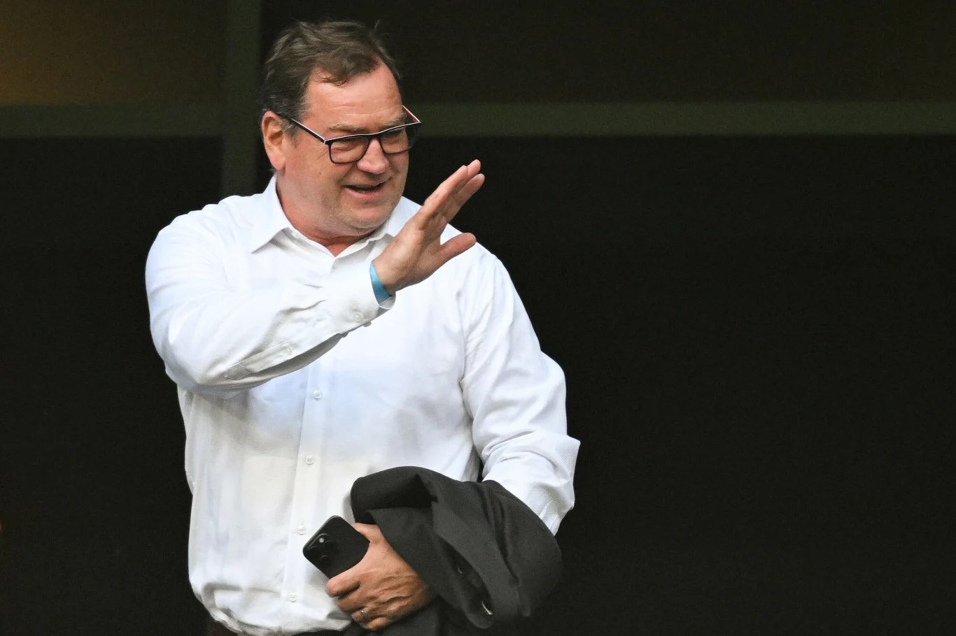 Jan Urban Uśmiechnięty mężczyzna w okularach i białej koszuli, trzymający marynarkę w jednej ręce i smartfon w drugiej, wykonuje gest powitania lub pożegnania, znajduje się na ciemnym tle.