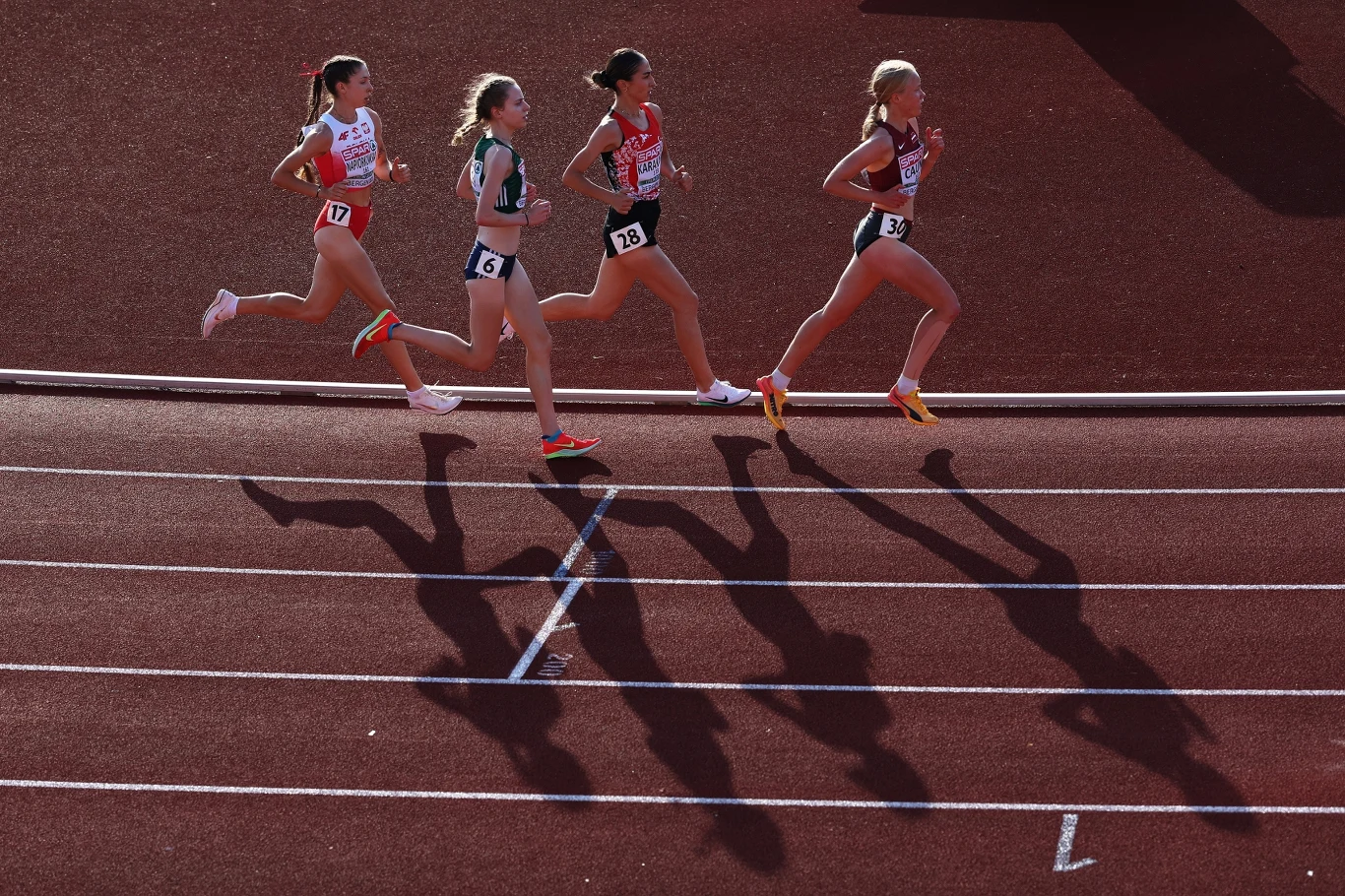 Katarzyna Napiórkowska (z lewej) przeżyła dramat na przedostatnim okrążeniu w biegu na 10000 metrów w ME U-23 w Bergen Cztery zawodniczki biegną na stadionowej bieżni, każda z przypiętym numerem startowym, ich sylwetki rzucają wyraziste cienie na podłoże, promienie słoneczne podkreślają dynamikę ruchu.