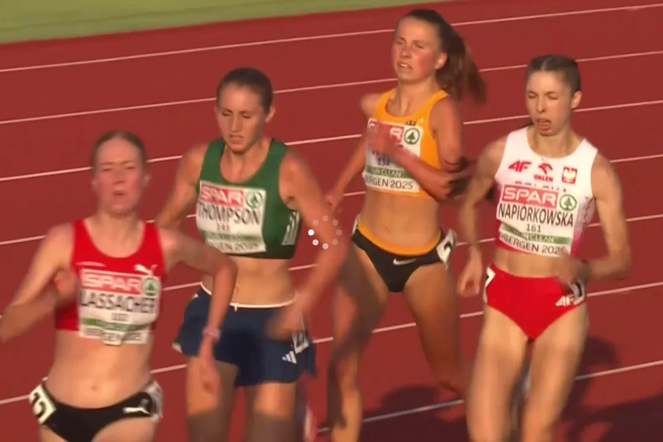 Moment dublowania Polki przez Irlandkę i Niemkę Grupa czterech lekkoatletek biegnących na stadionowej bieżni, z widocznymi tabliczkami startowymi i oznaczeniem zawodów, każda zawodniczka w stroju reprezentacyjnym swojego kraju, w trakcie intensywnej rywalizacji sportowej.