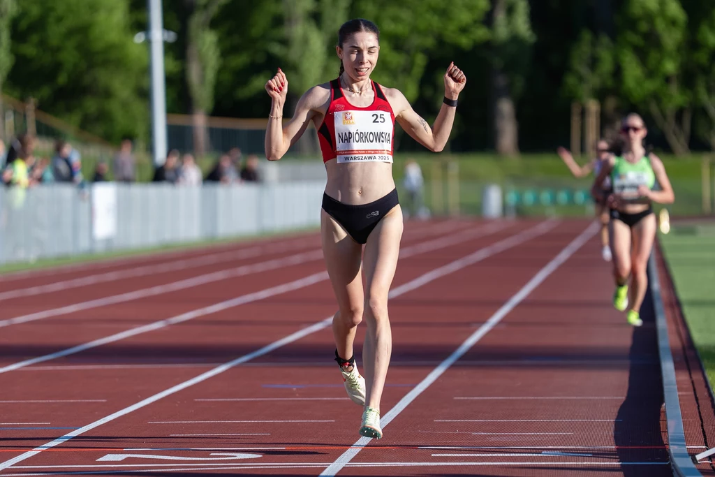 Zawodniczka biegnąca po bieżni lekkoatletycznej, unosi ręce w geście triumfu tuż po przekroczeniu mety, za nią w oddali widoczne są inne biegaczki oraz tło z zielenią i kibicami.
