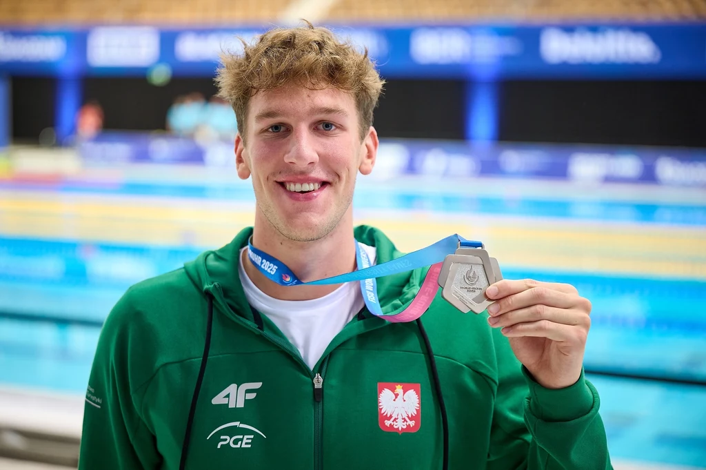 Młody mężczyzna w zielonej bluzie z godłem Polski oraz logo sponsora, uśmiechnięty, trzyma srebrny medal olimpijski na tle pływalni.