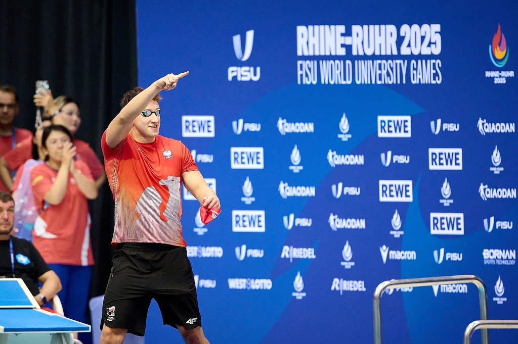Zawodnik w czerwonej koszulce i czarnych szortach przygotowuje się do gry w tenisa stołowego podczas zawodów, dynamicznie gestykulując rakietką, w tle niebieska plansza z logo FISU World University Games i napisami sponsorów.