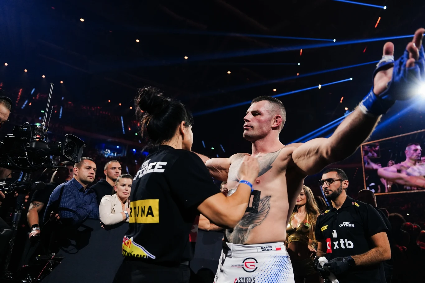 KSW: Marcin Wójcik Zawodnik MMA przygotowujący się do walki, rozkłada ramiona, podczas gdy członek zespołu sprawdza go przy wejściu do oktagonu. W tle publiczność, kamera i obsługa wydarzenia, oświetlone arenową scenografią.