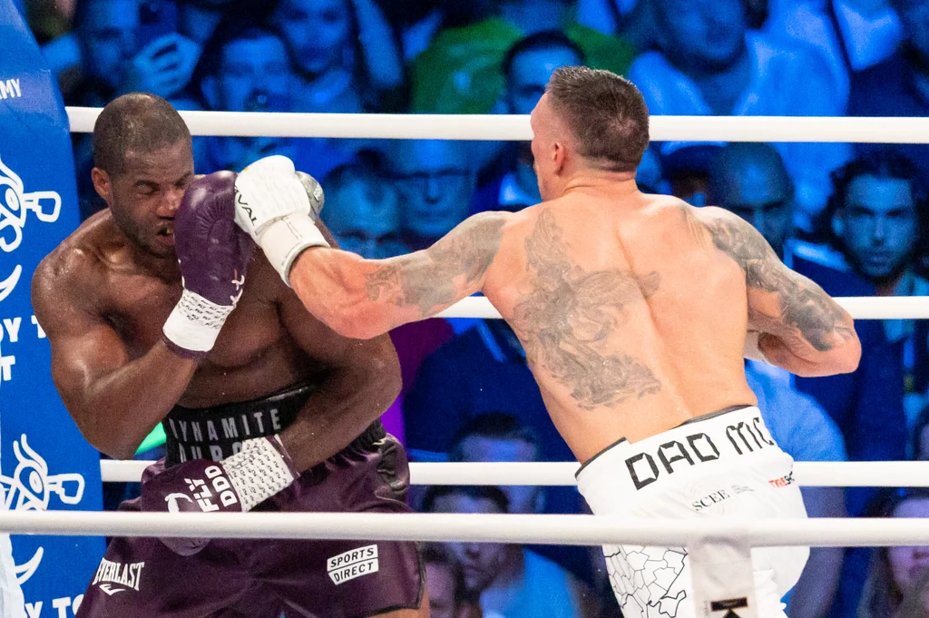 Daniel Dubois - Ołeksandr Usyk Dwóch bokserów w ringu podczas intensywnej walki, jeden z pięściarzy zadaje mocny cios przeciwnikowi w nocy, a tłum w tle emocjonalnie śledzi pojedynek.