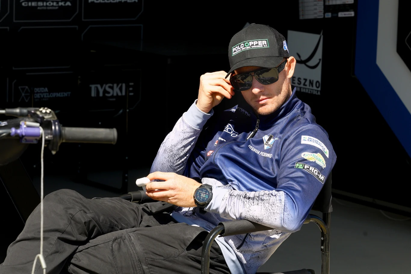 Josh Pickering na wylocie z Unii Leszno Mężczyzna w sportowej kurtce i czapce z daszkiem siedzi na krześle, trzymając okulary przeciwsłoneczne i patrzy w stronę aparatu, w tle widoczne są elementy techniczne oraz reklamy sponsorów.