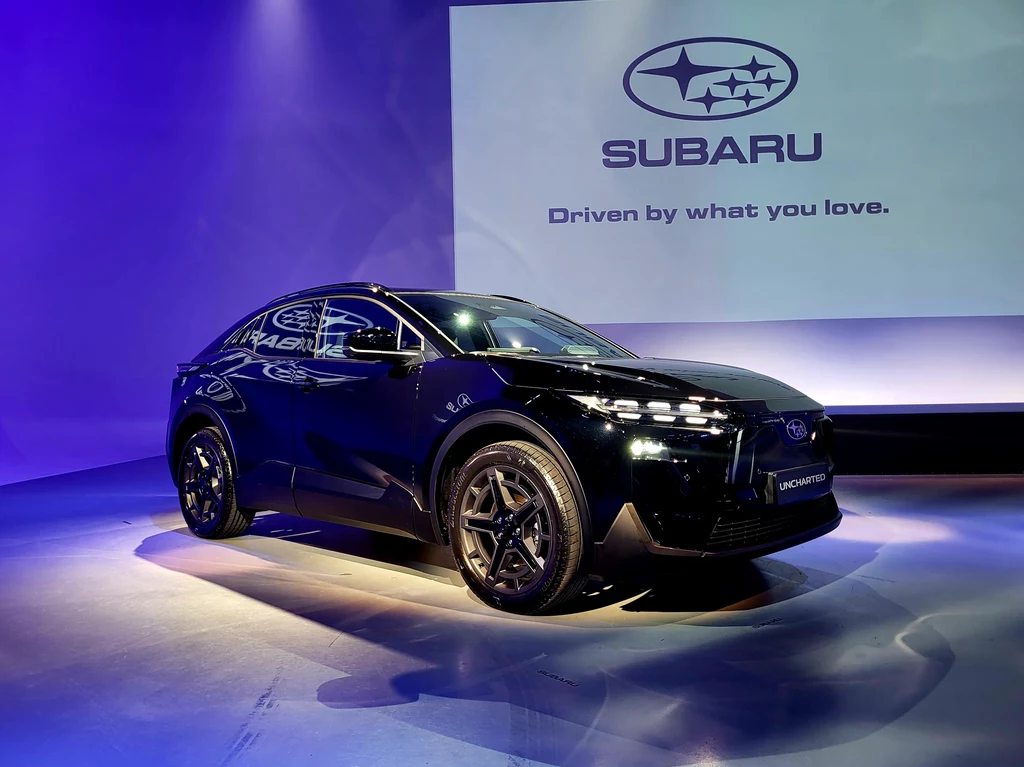 Nowoczesny czarny samochód Subaru ustawiony na tle fioletowego światła, z dużym podświetlonym logo Subaru oraz napisem „Driven by what you love” na ekranie w tle, scena o charakterze prezentacyjnym.