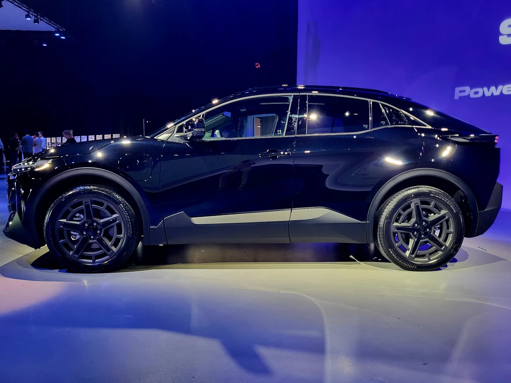 Czarny nowoczesny samochód elektryczny SUV sfotografowany z boku na tle oświetlonej sceny wystawowej, odbicia świateł na karoserii podkreślają elegancki design i aerodynamiczne kształty pojazdu.