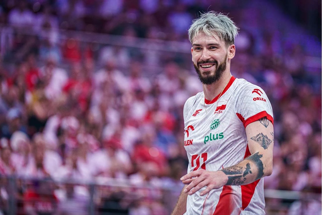 Siatkarz w białoczerwonym stroju z widocznymi tatuażami na rękach uśmiecha się podczas meczu, za nim widać zapełnione trybuny kibiców.