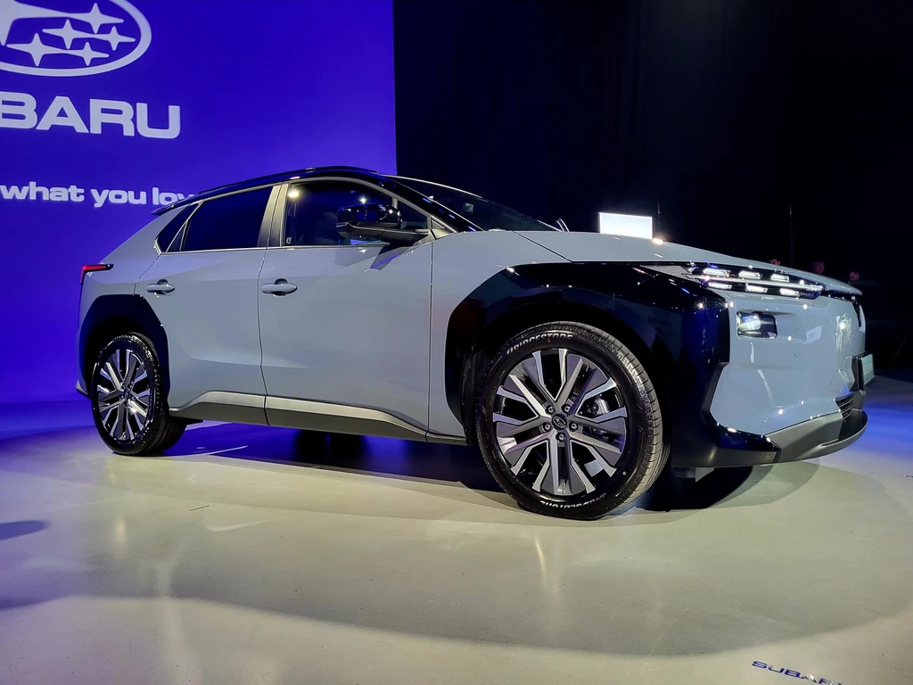 Nowoczesny samochód marki Subaru w jasnoszarym lakierze zaprezentowany na tle niebieskiego ekranu z logo i hasłem marki, podkreślone oświetleniem, zwracające uwagę na detale stylistyczne i futurystyczny wygląd pojazdu.