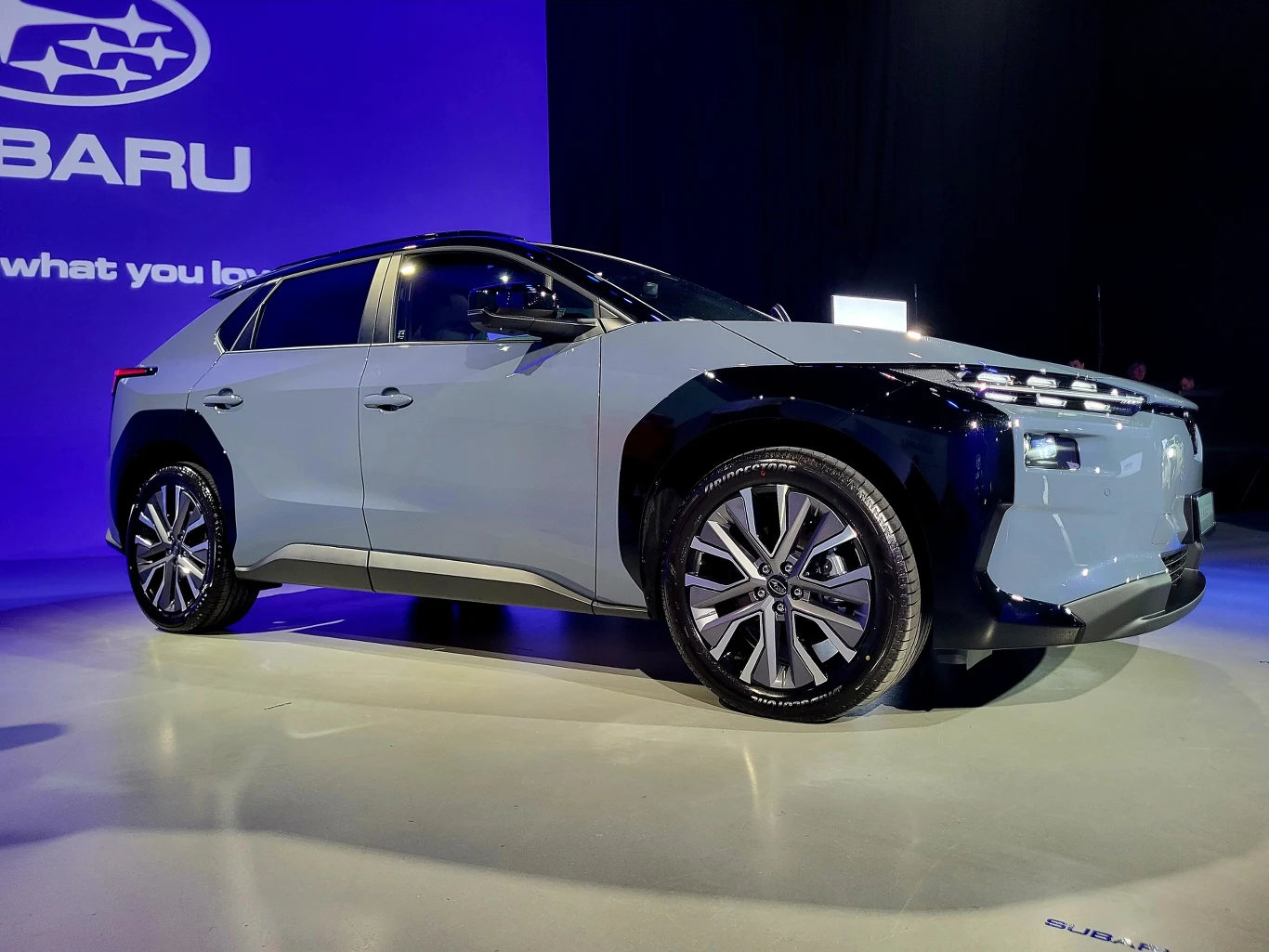 Nowoczesny samochód marki Subaru w jasnoszarym lakierze zaprezentowany na tle niebieskiego ekranu z logo i hasłem marki, podkreślone oświetleniem, zwracające uwagę na detale stylistyczne i futurystyczny wygląd pojazdu.