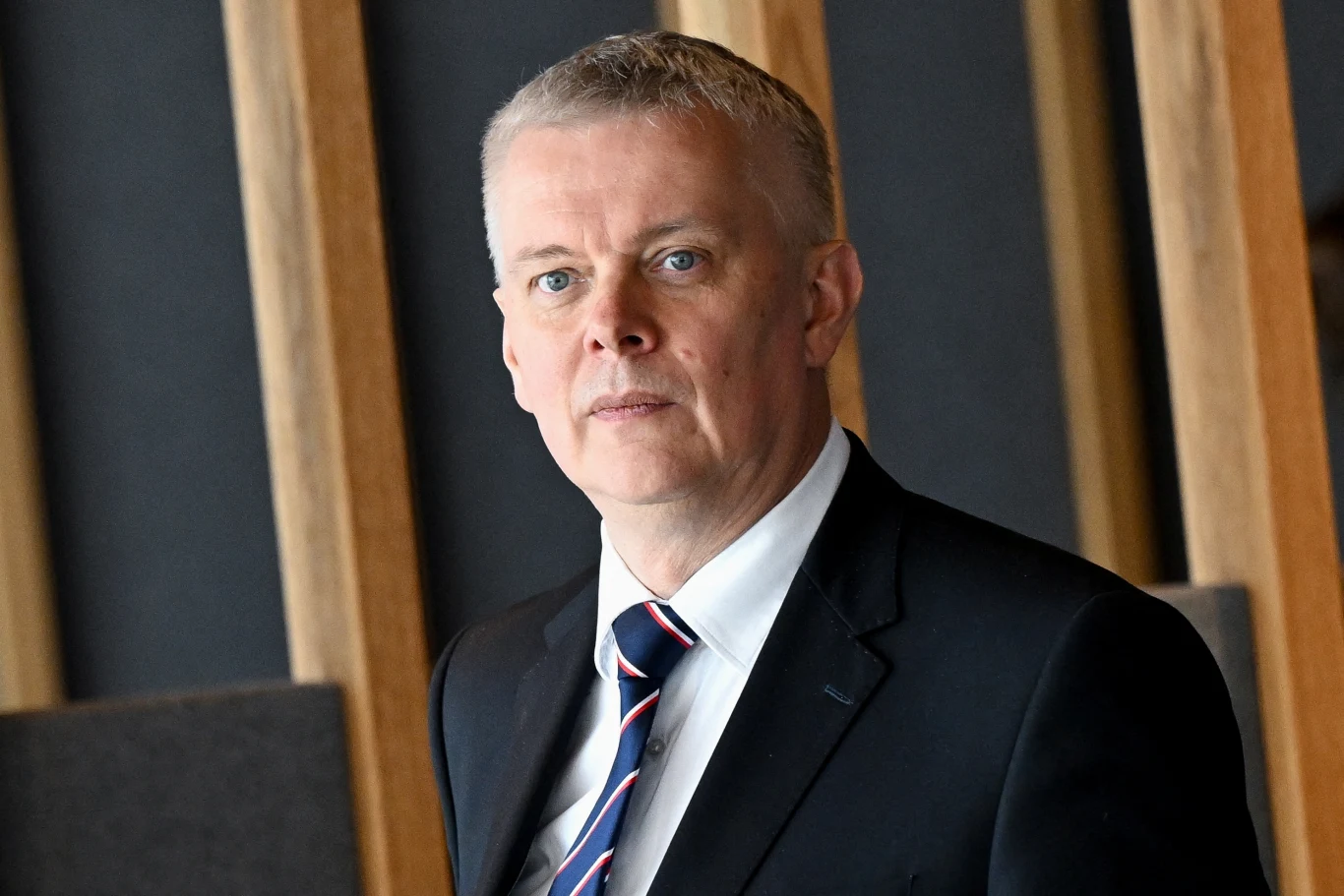 Awaria na lotniskach, natychmiastowa reakcja ministra. Służby uruchomione Mężczyzna w średnim wieku w garniturze z krawatem, krótkie siwe włosy, neutralna mina, patrzący wprost na fotografa, w tle drewno oraz szaro-czarna ściana.