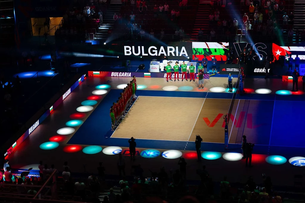 Uroczyste ustawienie dwóch drużyn siatkarskich na boisku podczas oficjalnej prezentacji przed meczem, na tle podświetlonego napisu z nazwą kraju Bułgaria i jej flagą.