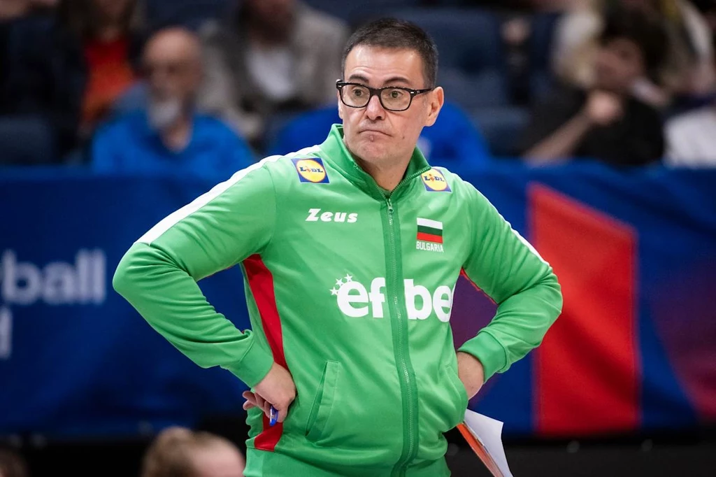 Mężczyzna w zielonej bluzie sportowej z flagą Bułgarii oraz logotypami sponsorów stoi na tle trybun, z poważnym wyrazem twarzy i założonymi rękami