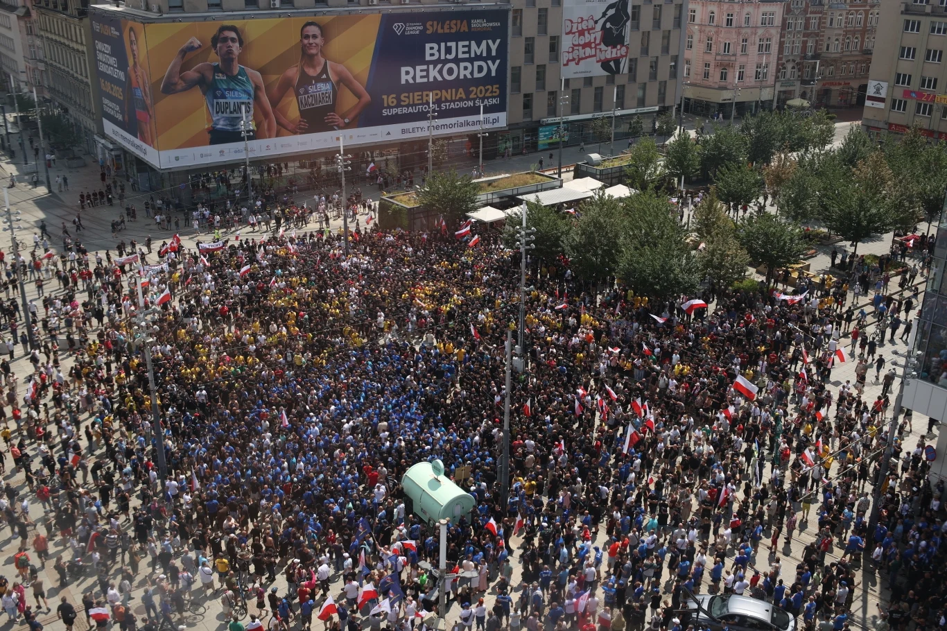 Protest przeciw nielegalnej migracji w Katowicach Tłum ludzi zgromadzonych na miejskim placu, część osób trzyma flagi narodowe, widoczna scena oraz bilbord promujący wydarzenie sportowe, wokół drzewa i miejskie zabudowania.