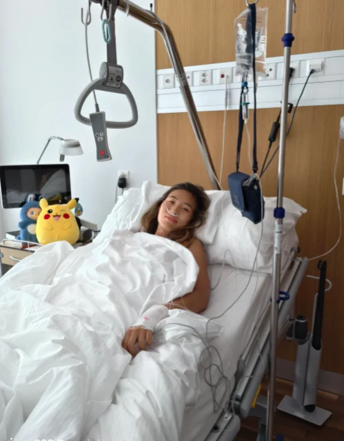 Kobieta leży w szpitalnym łóżku, otulona białą pościelą, otoczona medycznym sprzętem i kroplówkami, obok na szafce znajduje się pluszowa zabawka Pikachu.