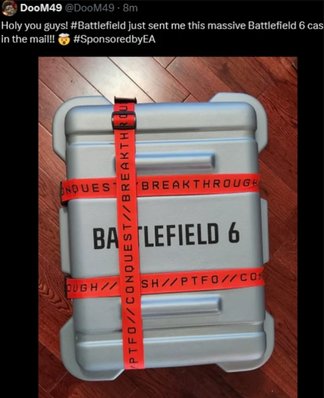 Duża, metalowa skrzynka z napisem 'Battlefield 6', opasana czerwoną taśmą z nadrukami 'CONQUEST', 'BREAKTHROUGH', 'PTFO'. Skrzynka stoi na drewnianej podłodze. Na zdjęciu widoczny jest również wpis w mediach społecznościowych dotyczący otrzymania tej p...