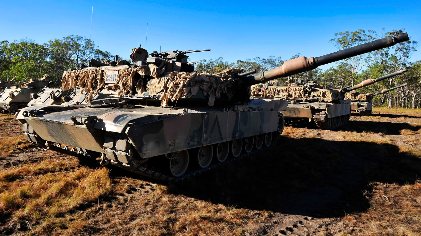 Czołg bojowy typu Abrams ustawiony na otwartej przestrzeni, zamaskowany siatką, w otoczeniu innego sprzętu pancernego; sucha roślinność i drzewa tworzą tło kadru.