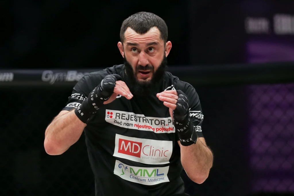 Mamed Chalidow Zawodnik MMA z brodą w czarnej koszulce z logotypami sponsorów stoi w ringu, trzymając gardę w pozycji obronnej i koncentrując wzrok na przeciwniku.