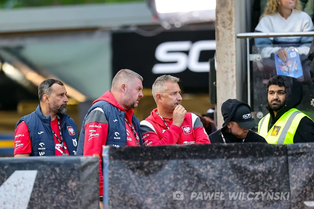 Sztab reprezentacji Polski Trzech mężczyzn w sportowej odzieży z emblematami Polski stoi na trybunach, koncentrując się na wydarzeniu, jeden z nich opiera rękę o usta w zamyśleniu, w tle widać ochroniarza i przeszkloną lożę z obserwatorami.