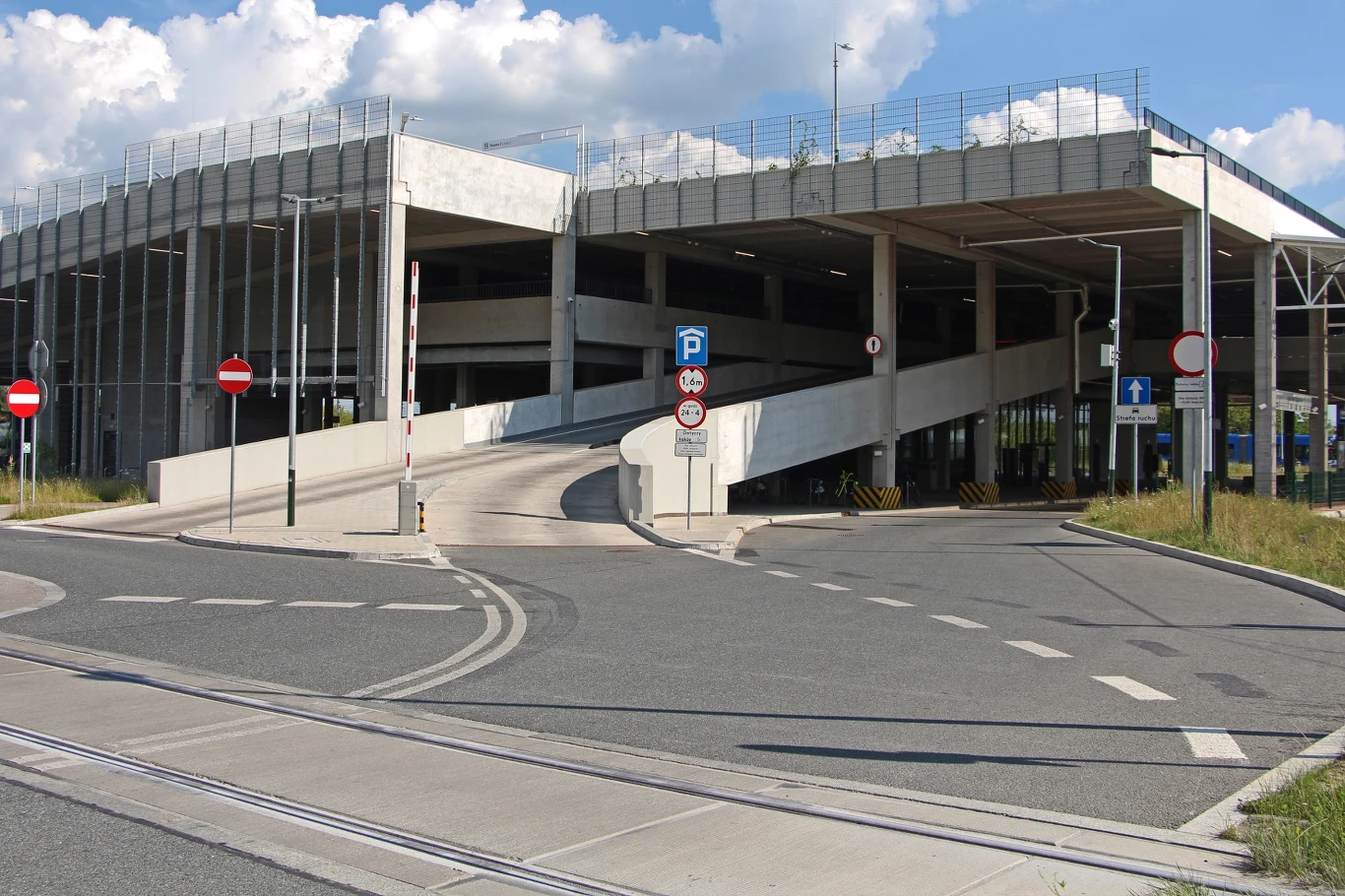 Parking P+R Górka Narodowa znajduje się tuż przy zjeździe z północnej obwodnicy Krakowa Wielopoziomowy parking z betonową rampą wjazdową, licznymi znakami drogowymi zakazu wjazdu i informującymi o parkingu, otoczony asfaltową drogą oraz fragmentem zieleni przy krawężniku, na tle nieba z chmurami.