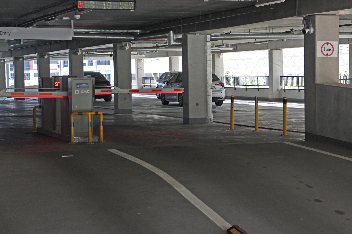 Również przed bramkami na wjeździe do parkingu nie znajdziemy żadnego fizycznego ograniczenia dla pojazdów mających ponad 1,6 m wysokości - tylko znak na filarze Parking podziemny z zamontowanymi szlabanami, kilka zaparkowanych samochodów, słupy podtrzymujące konstrukcję i oznaczenie maksymalnej wysokości wjazdu 1,9 m.