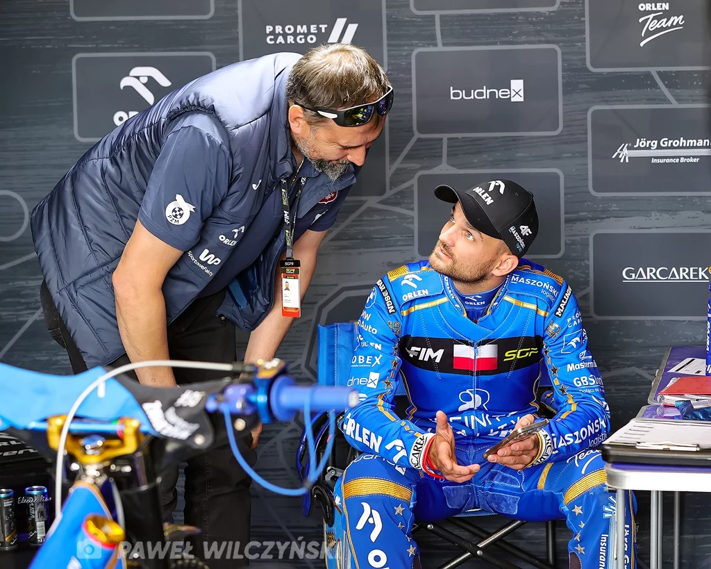 Rafał Dobrucki, Bartosz Zmarzlik Dwóch mężczyzn w sportowych ubraniach prowadzi rozmowę w boksie, jeden z nich siedzi w niebieskim kombinezonie motocyklowym z logotypami sponsorów, drugi stoi obok w kamizelce z identyfikatorem.