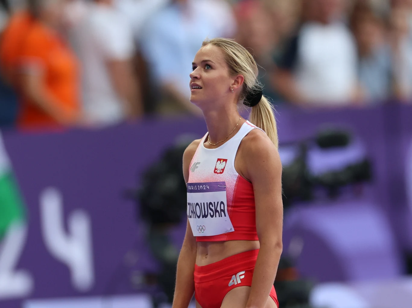 Sportowczyni w stroju reprezentacji Polski stoi na stadionie lekkoatletycznym, skupiona i gotowa do startu, w tle niewyraźni widzowie i elementy toru olimpijskiego.
