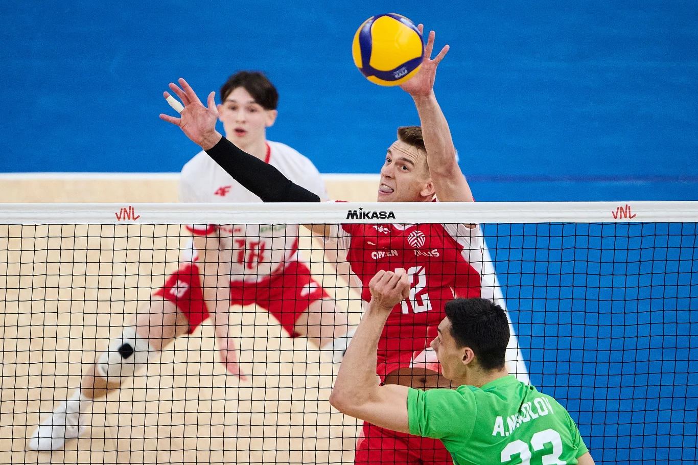 Artur Szalpuk w trakcie meczu Polska - Bułgaria Dynamiczna akcja podczas meczu siatkówki, jeden z zawodników w czerwonym stroju skacze, by zablokować piłkę nad siatką, po drugiej stronie gracz w zielonej koszulce próbuje zdobyć punkt, a w tle widoczni są inni członkowie drużyn.