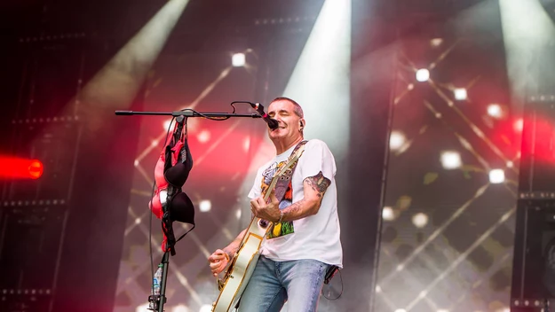Wokalista z gitarą elektryczną występuje na scenie podczas koncertu muzycznego, w tle kolorowe światła, na statywie mikrofonu zawieszony biustonosz, widoczny fragment drugiego muzyka.
