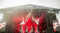 Duża scena koncertowa z licznymi reflektorami i efektami świetlnymi o czerwonej barwie, muzycy występujący na scenie, tłum ludzi obserwujących wydarzenie na pierwszym planie, widoczne logo festiwalu Jarocin Festiwal oraz daty wydarzenia.