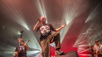 Zespół muzyczny występujący na scenie podczas koncertu, wokalista śpiewa do mikrofonu z energiczną pozą, wokół muzyków efektowne światła sceniczne podkreślają dynamiczną atmosferę wydarzenia.