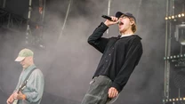 Dwóch muzyków na scenie podczas koncertu, wokalista dynamicznie śpiewa do mikrofonu, gitarzysta skupiony na grze; w tle widoczny dym sceniczny oraz elementy nagłośnienia i oświetlenia.