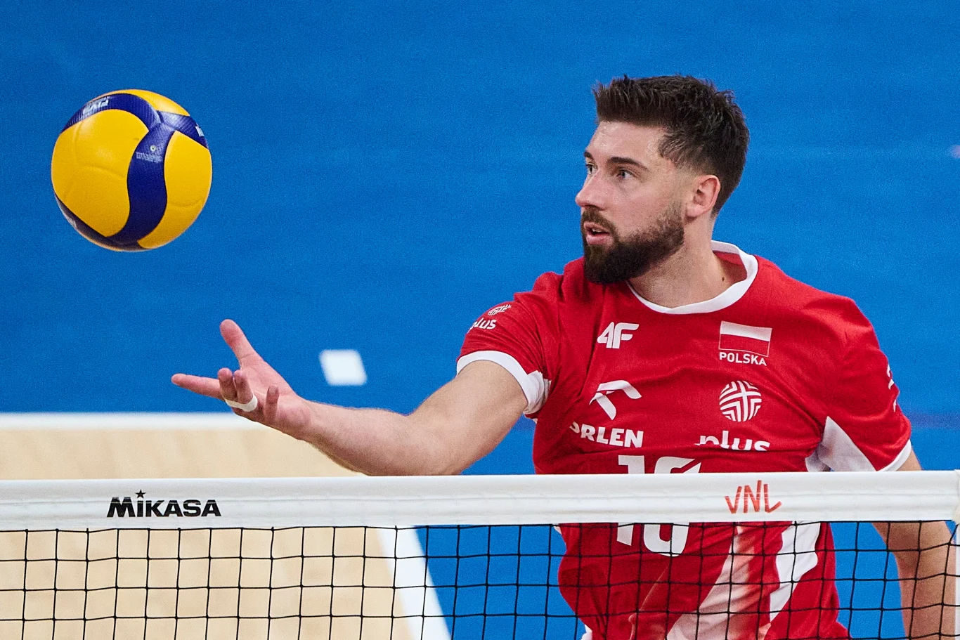 Bartosz Bednorz Polski siatkarz w czerwonej koszulce wykonuje serwis ponad siatką podczas meczu siatkówki, na pierwszym planie piłka do gry oraz fragment boiska.