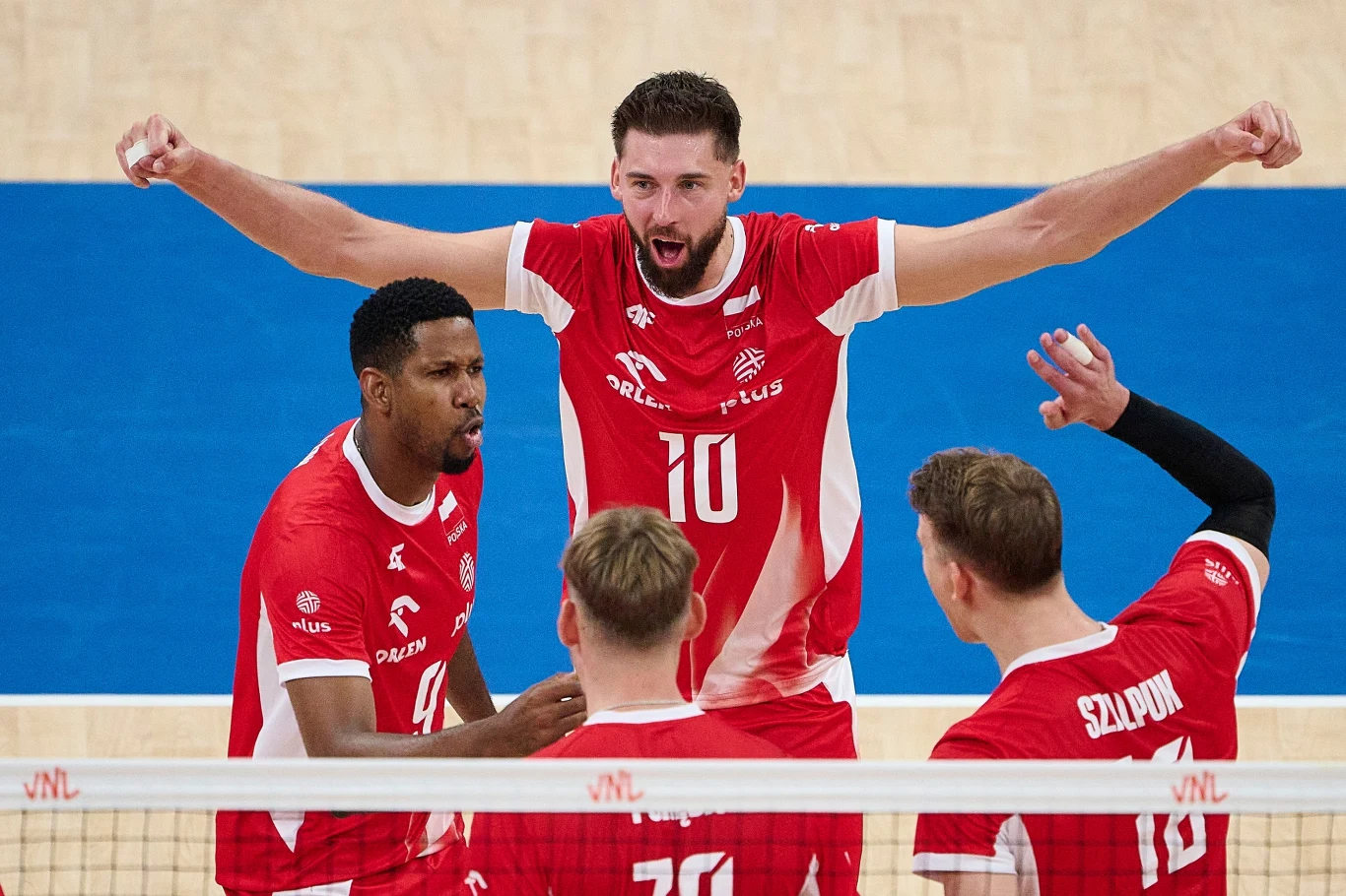 Bartosz Bednorz w centralnym punkcie fotografii Grupa siatkarzy w czerwonych strojach świętuje zdobycie punktu, jeden z zawodników stoi z uniesionymi ramionami, pozostali patrzą w jego stronę, widoczna siatka do gry i fragment boiska.