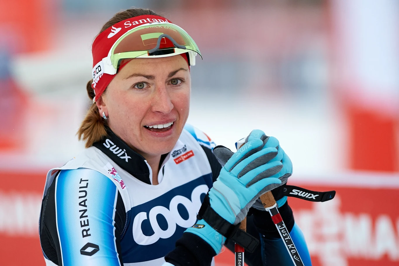 zawodniczka narciarstwa biegowego w stroju sportowym, trzymająca kijki, z czapką i okularami na głowie, widoczna w tle infrastruktura sportowa