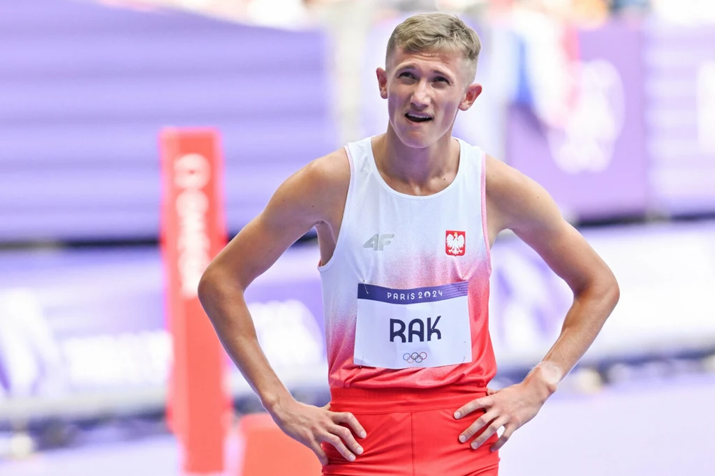 Filip Rak Lekkoatleta w stroju reprezentacji Polski, z rękami na biodrach, na tle bieżni sportowej i rozmytych elementów hali sportowej.