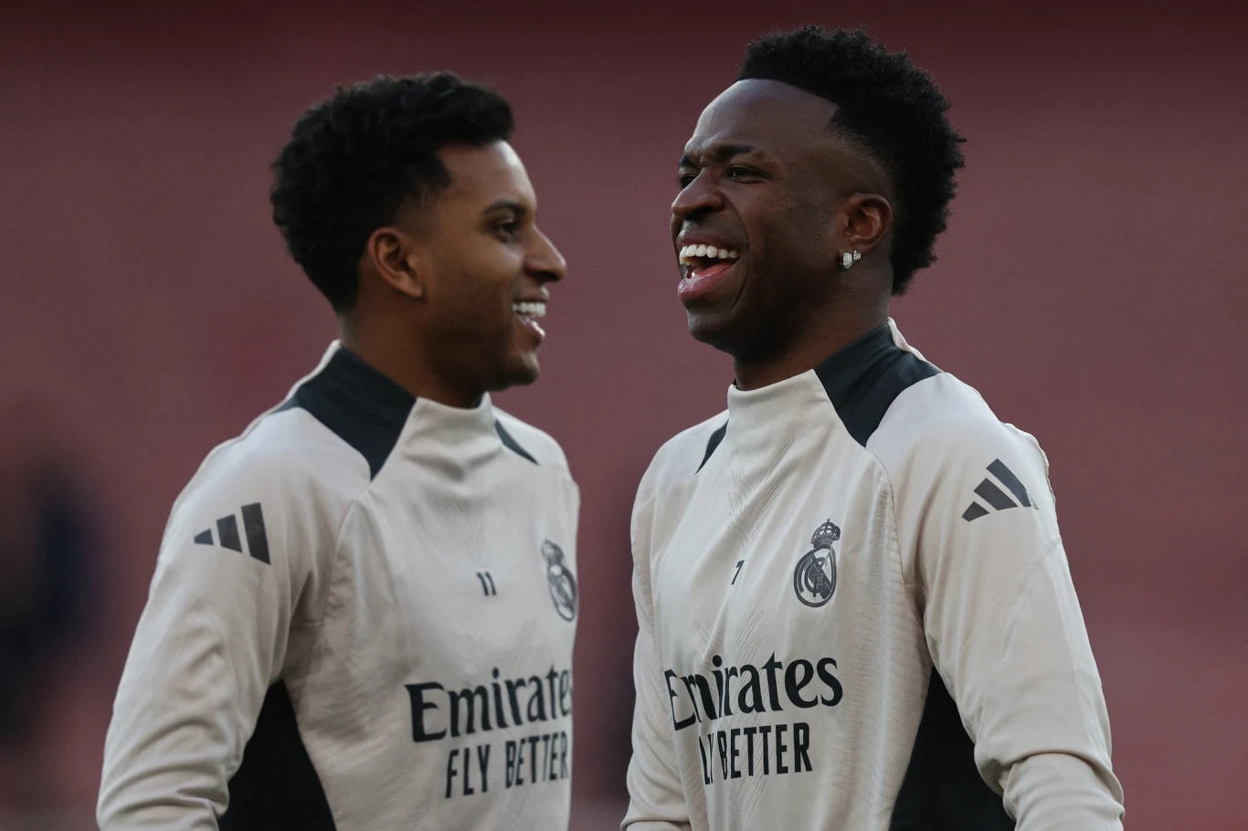 Dwóch piłkarzy uśmiecha się i rozmawia ze sobą podczas treningu, ubrani w jasne stroje sportowe drużyny Real Madryt na tle jednokolorowego tła.