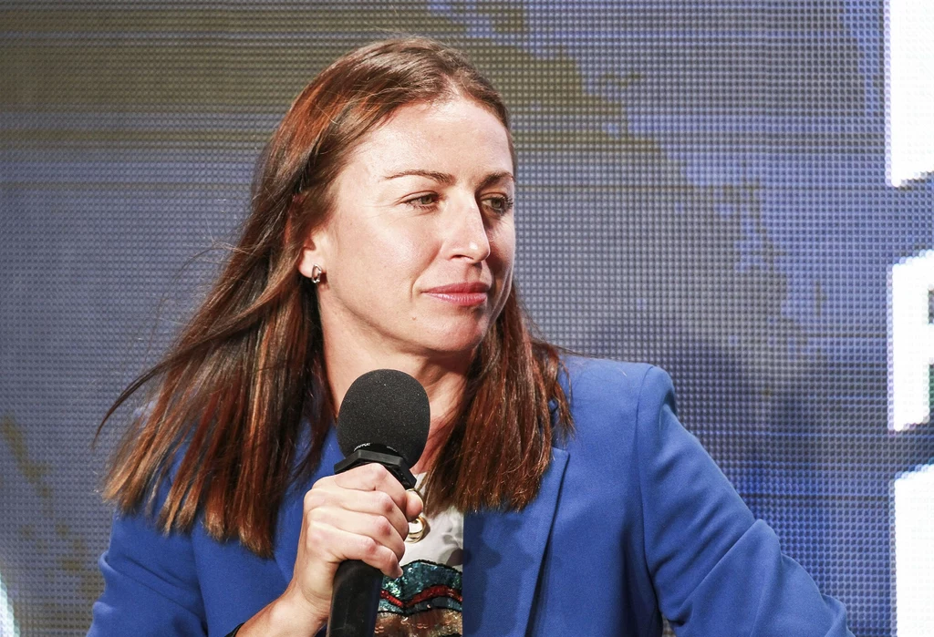 Justyna Kowalczyk-Tekieli Kobieta w średnim wieku trzyma mikrofon, ubrana w niebieską marynarkę, z długimi brązowymi włosami, siedzi na tle ekranu z elementami graficznymi.