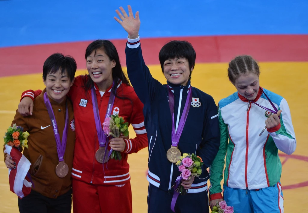 Hitomi Obara w Londynie zdobyła upragniony złoty medal Igrzysk Olimpijskich Cztery zawodniczki stoją obok siebie z medalami na szyjach i bukietami kwiatów, ubrane w stroje reprezentacyjne różnych krajów, jedna z nich uśmiecha się szeroko i macha ręką w kierunku publiczności.