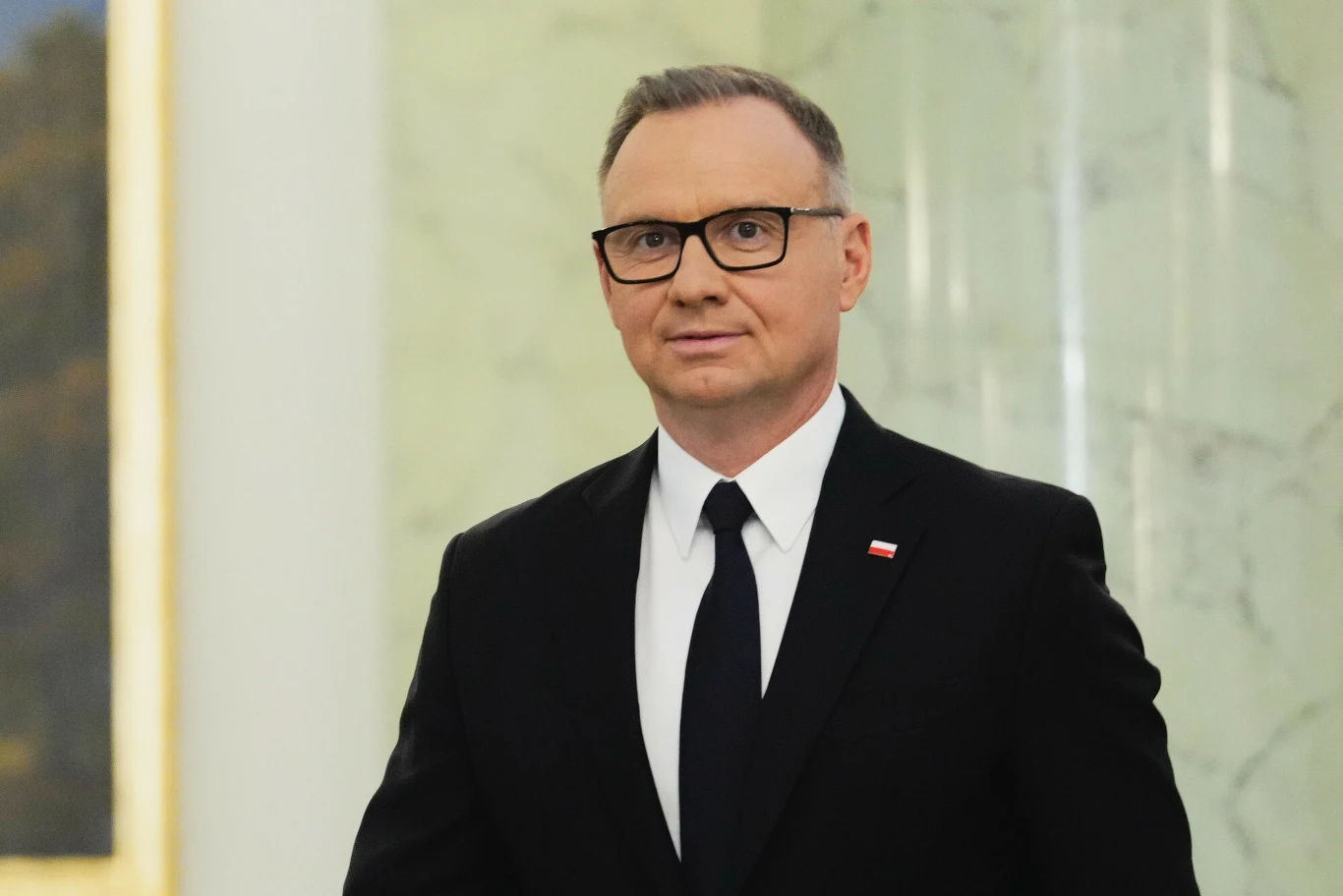 Polacy ocenili ostatnie wypowiedzi prezydenta Andrzeja Dudy Mężczyzna w okularach w ciemnym garniturze, białej koszuli i czarnej krawacie stoi na tle jasnej ściany z delikatnym wzorem marmuru, przypinka z flagą Polski widoczna na klapie marynarki