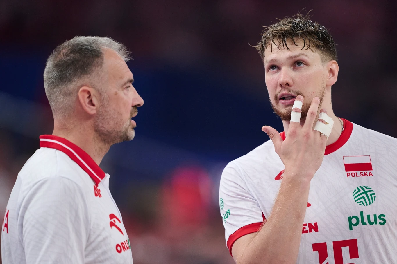 Zbliżenie dwóch mężczyzn w sportowych koszulkach reprezentacji Polski siatkówki, jeden z nich z opatrzonym palcem na dłoni gestykuluje podczas rozmowy, drugi uważnie słucha.