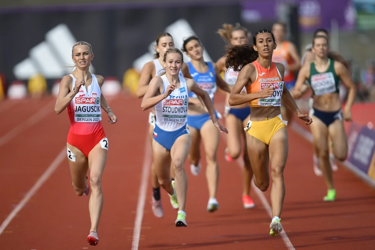 Grupa kobiet biegnących na bieżni stadionu lekkoatletycznego podczas zawodów, wszystkie w strojach reprezentacyjnych swoich krajów, z wyraźnie widocznymi numerami startowymi i nazwiskami.