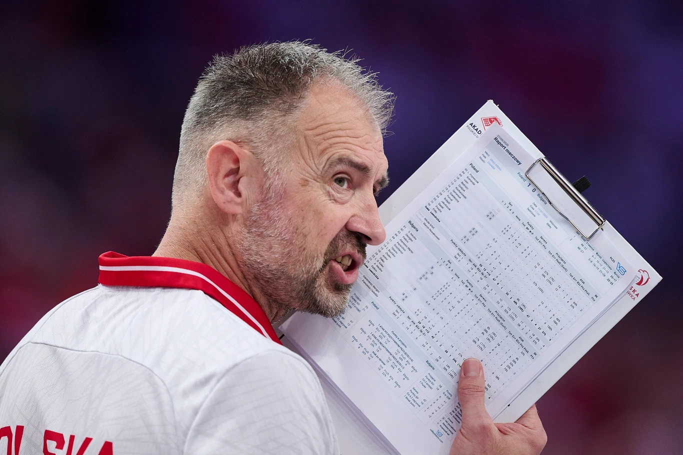 Nikola Grbić zachowuje spokój Mężczyzna w sportowej koszulce z napisem Polska trzyma w ręku tablicę z notatkami i arkuszami, odwrócony bokiem w kierunku kamery, w tle rozmyte kolory i światło.