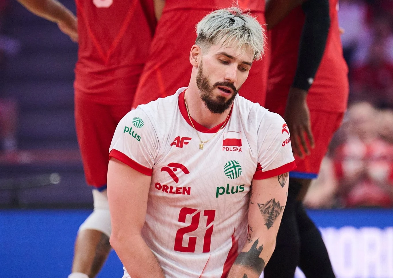 Zawodnik siatkarski w białoczerwonym stroju reprezentacji Polski z numerem 21, z blond włosami i tatuażem na lewym ramieniu, wygląda na zmartwionego lub zmęczonego, w tle widoczni inni siatkarze w czerwonych strojach.