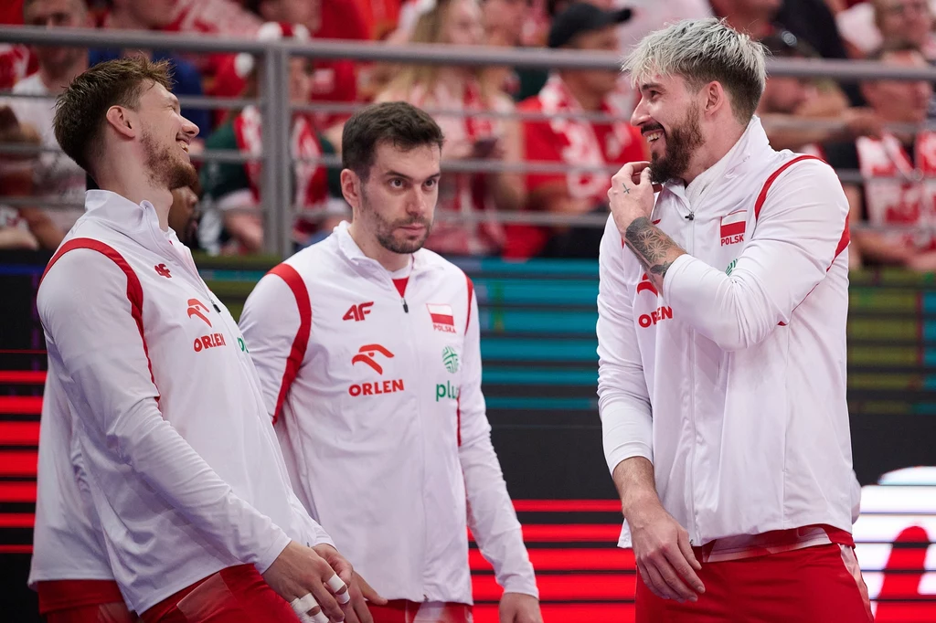 Tomasz Fornal (z prawej) Trzech siatkarzy reprezentacji Polski ubranych w biało-czerwone stroje rozmawia i żartuje ze sobą na tle trybun wypełnionych kibicami.