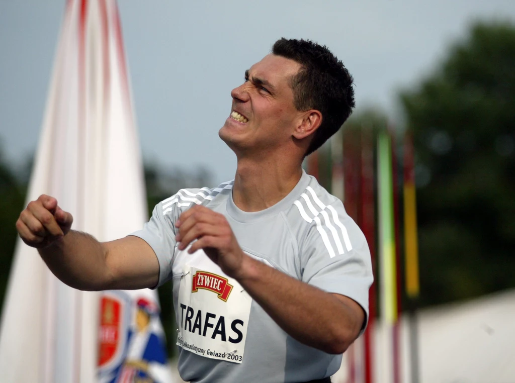 Zawodnik w sportowej koszulce z identyfikatorem wykonuje dynamiczny gest, jego twarz wyraża skupienie i wysiłek, w tle widoczne są rozmyte flagi i elementy stadionu.