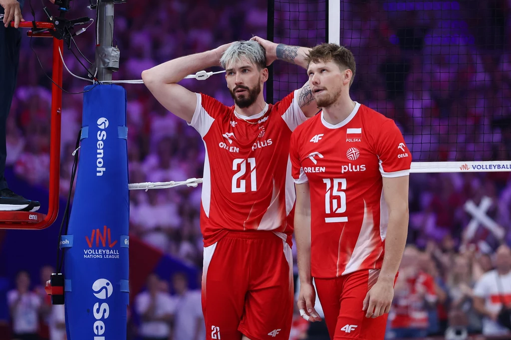 Tomasz Fornal (z lewej) i Jakub Kochanowski Dwóch siatkarzy w czerwonych strojach reprezentacji Polski stoi obok siebie na boisku, jeden z nich trzyma ręce na głowie, wyraz twarzy sugeruje skupienie lub zaniepokojenie, w tle widoczna rozmazywana publiczność i elementy hali sportowej.
